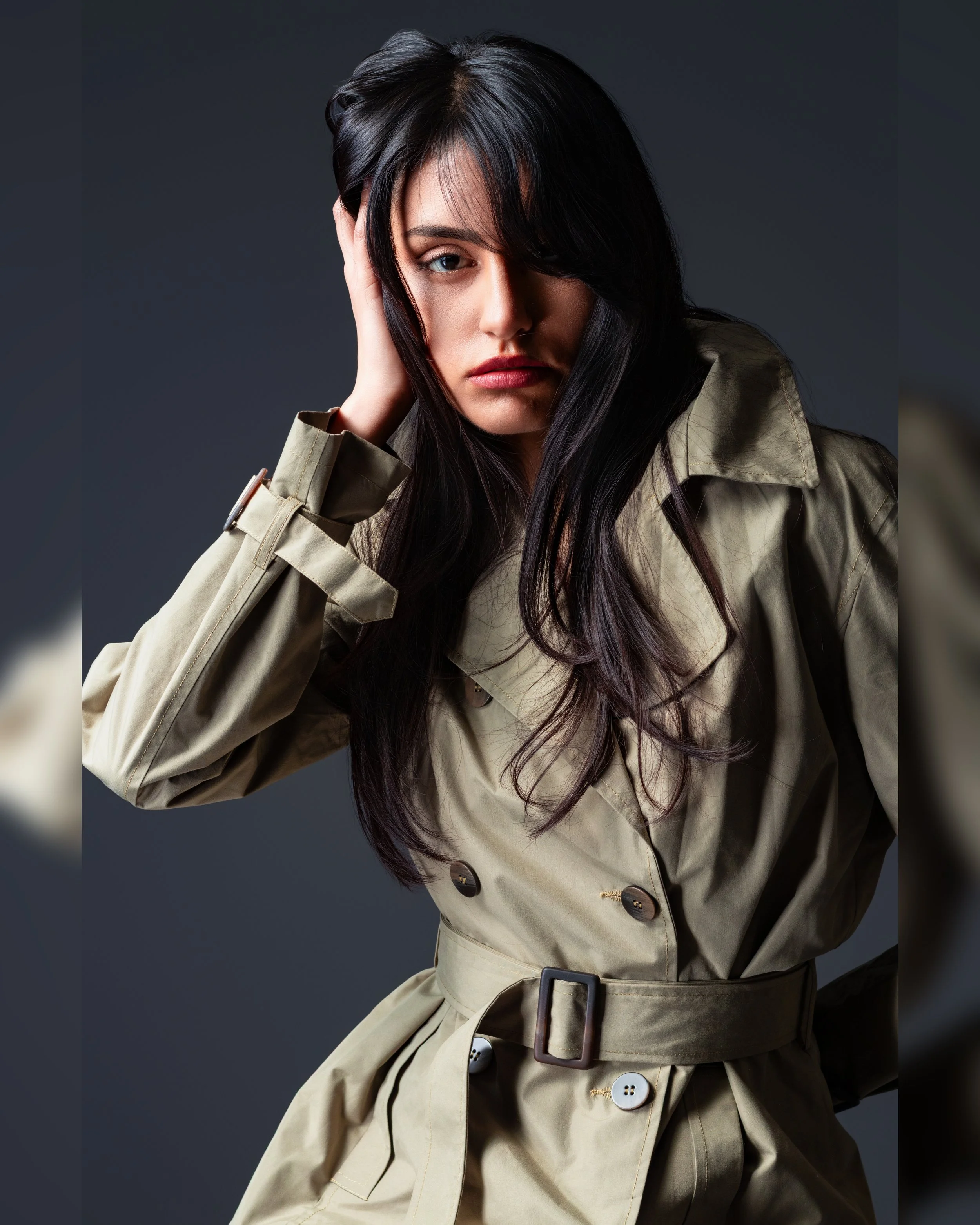 A woman with long dark hair, fair skin, and blue eyes, wearing a beige trench coat with a belt, resting her head on her hand against a dark gray background.