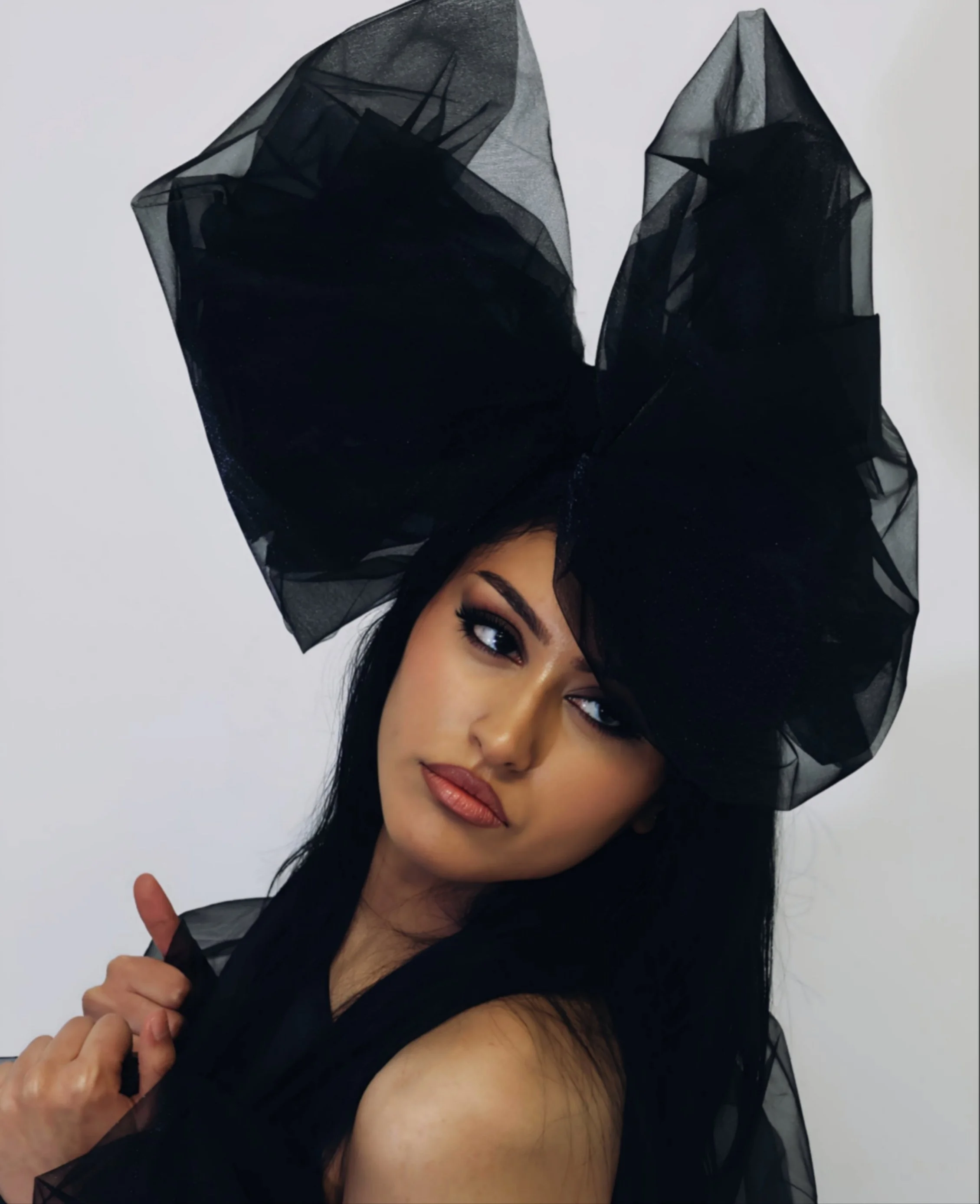 A woman with dark hair and makeup wearing a black sheer fabric headpiece with structured wings and a black garment, holding a black tie or fabric strip, against a plain light background.