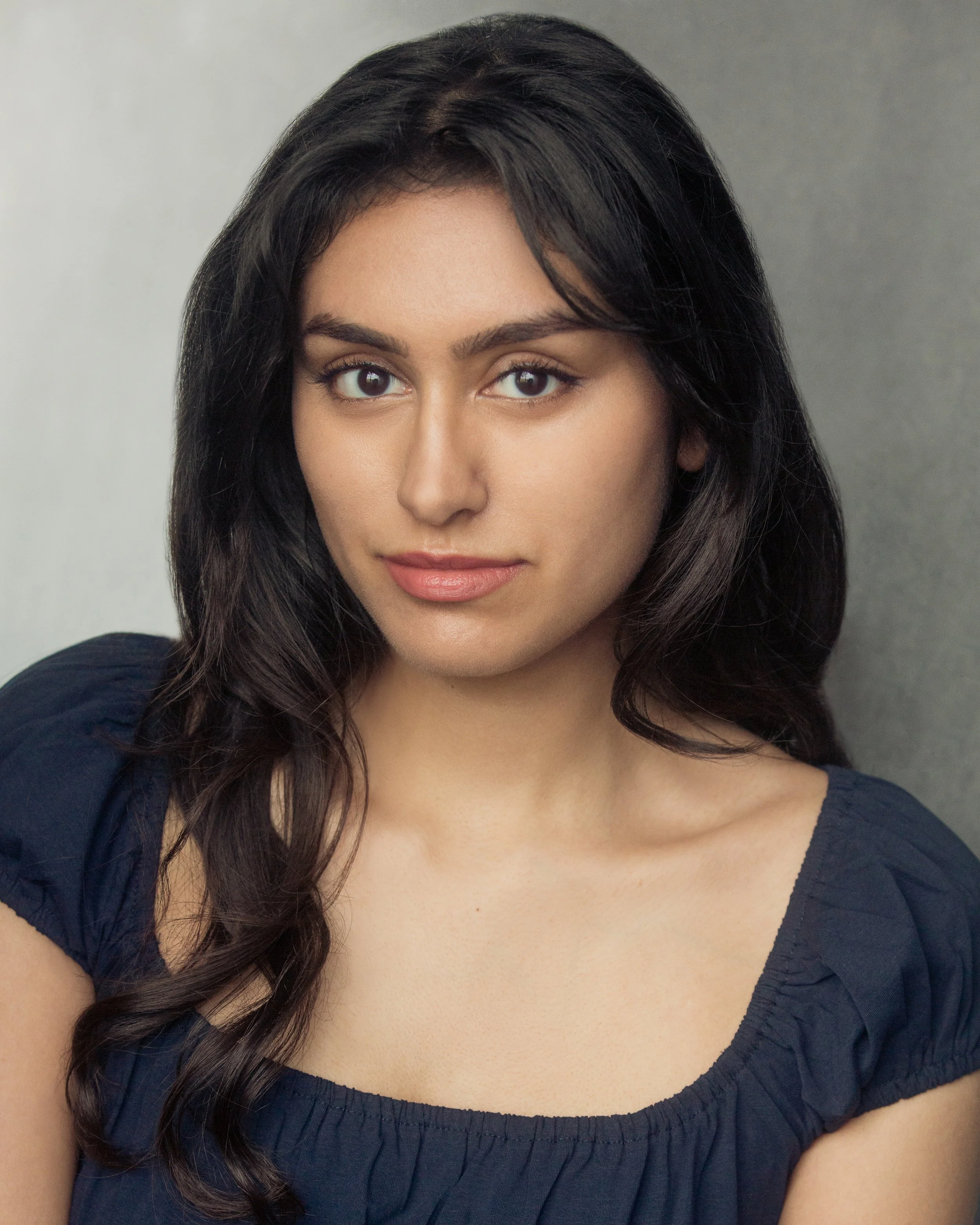 A woman with long, dark hair wearing a navy blue top with a ruffled neckline, smiling gently at the camera against a neutral background.