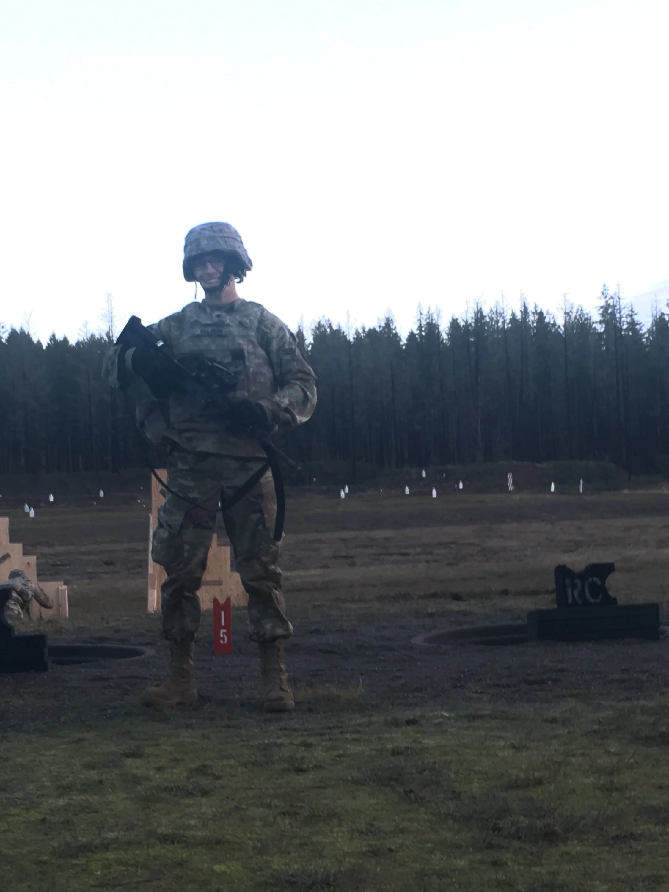 A soldier in camouflage uniform and helmet holding a firearm, standing outdoors at a training or shooting range with markers and targets in the background.