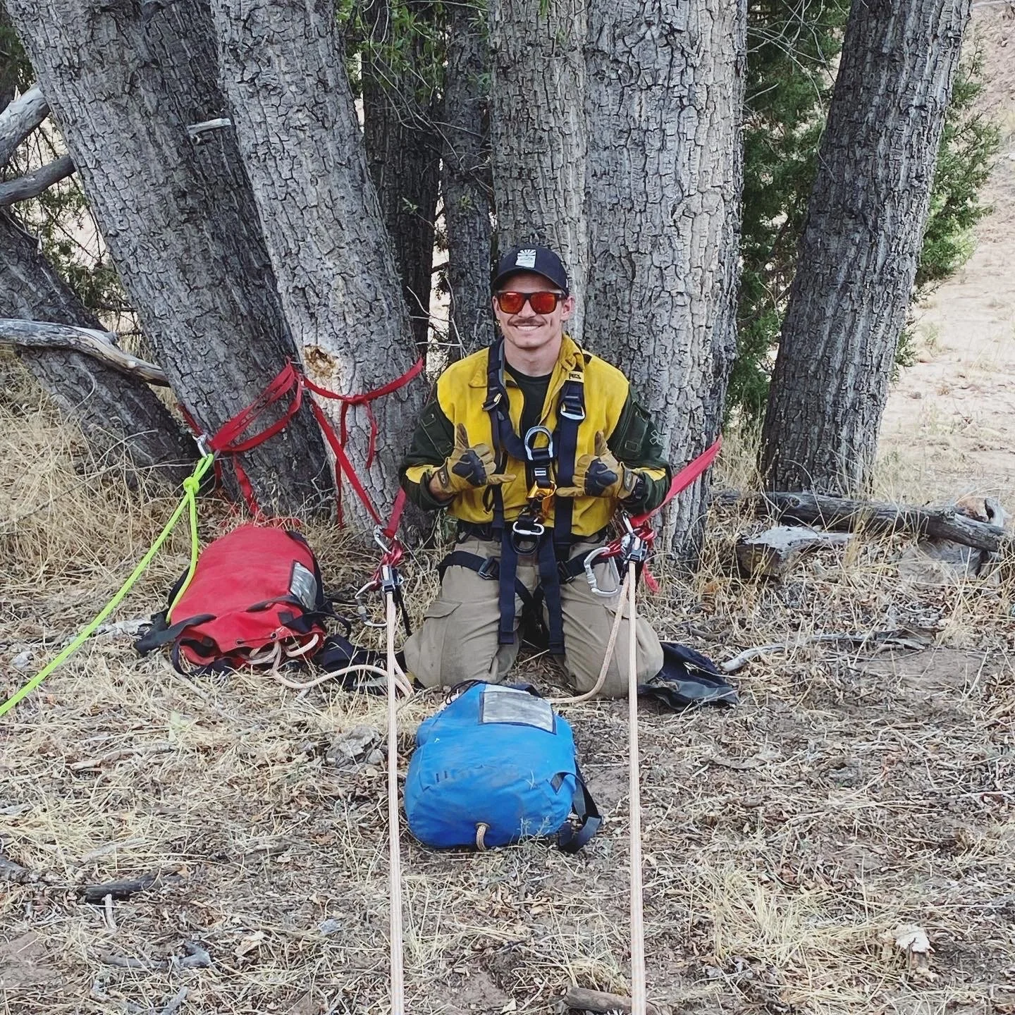 A man kneeling on the ground in front of a large tree, smiling and giving thumbs-up, surrounded by tents and camping gear in a dry, grassy outdoor area. Rope Rescue/ REMs