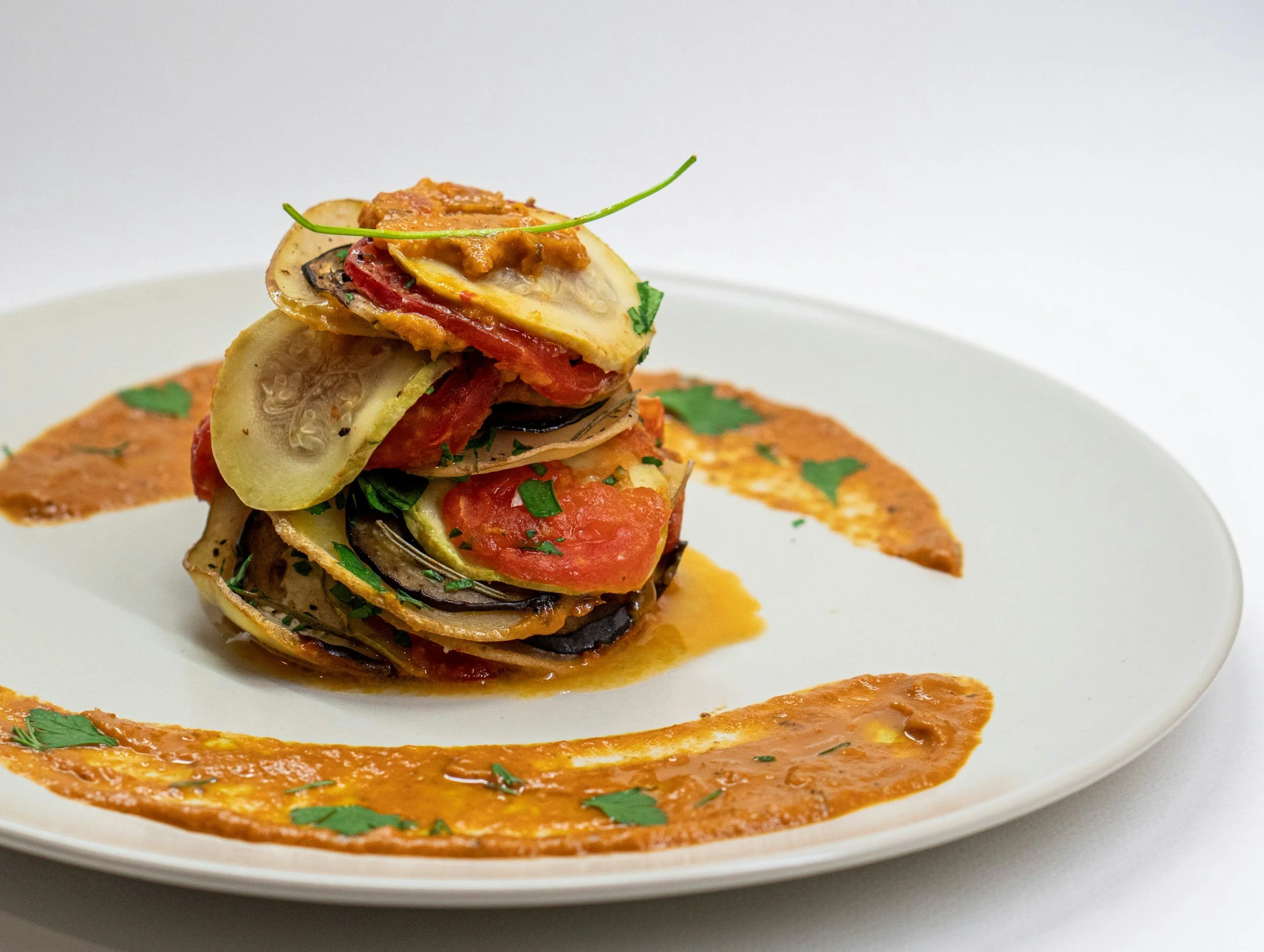 Stacked grilled vegetables and tomato slices garnished with a green herb on a white plate with a red sauce background.