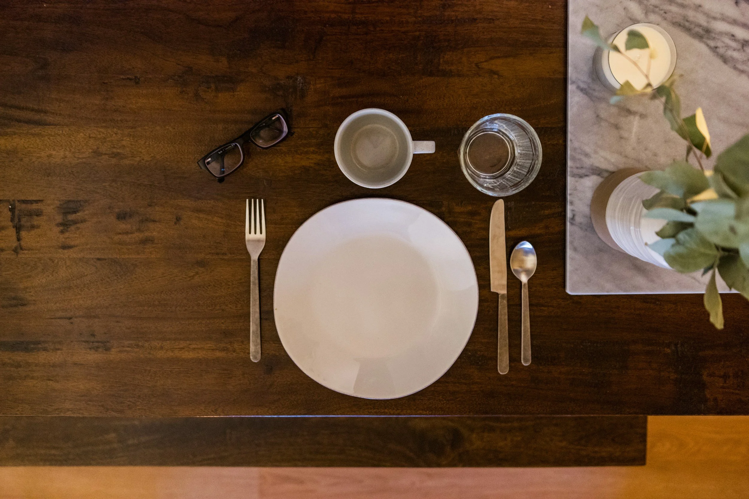 A wooden dining table set up for a meal with a white plate, a fork, a knife, a spoon, a clear glass of water, a white mug, a pair of glasses, and a vase with green leaves and yellow flowers on a marble surface.