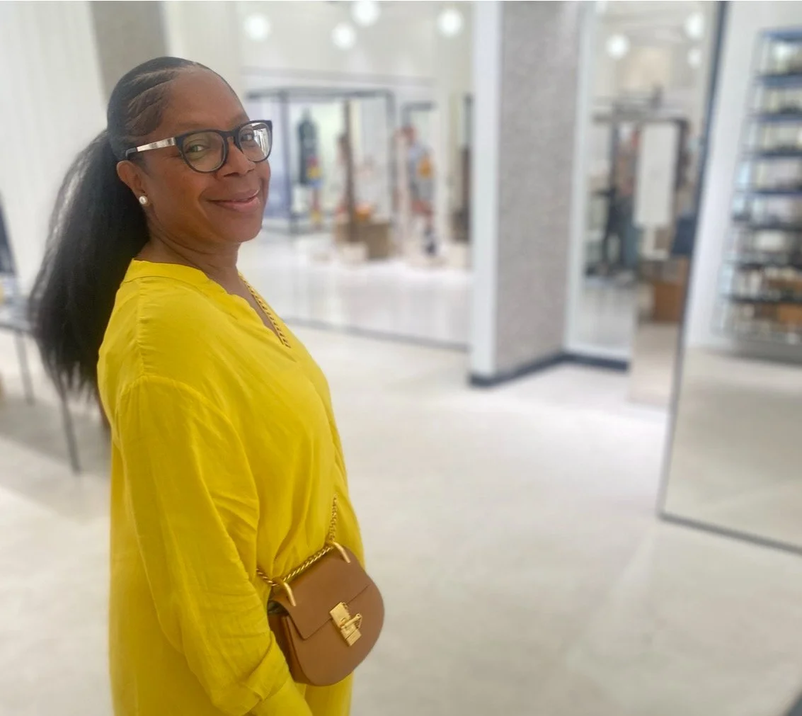 A woman with glasses and earrings wearing a yellow blouse and carrying a small brown purse, standing in a modern indoor space with bookshelves and glass doors in the background.
