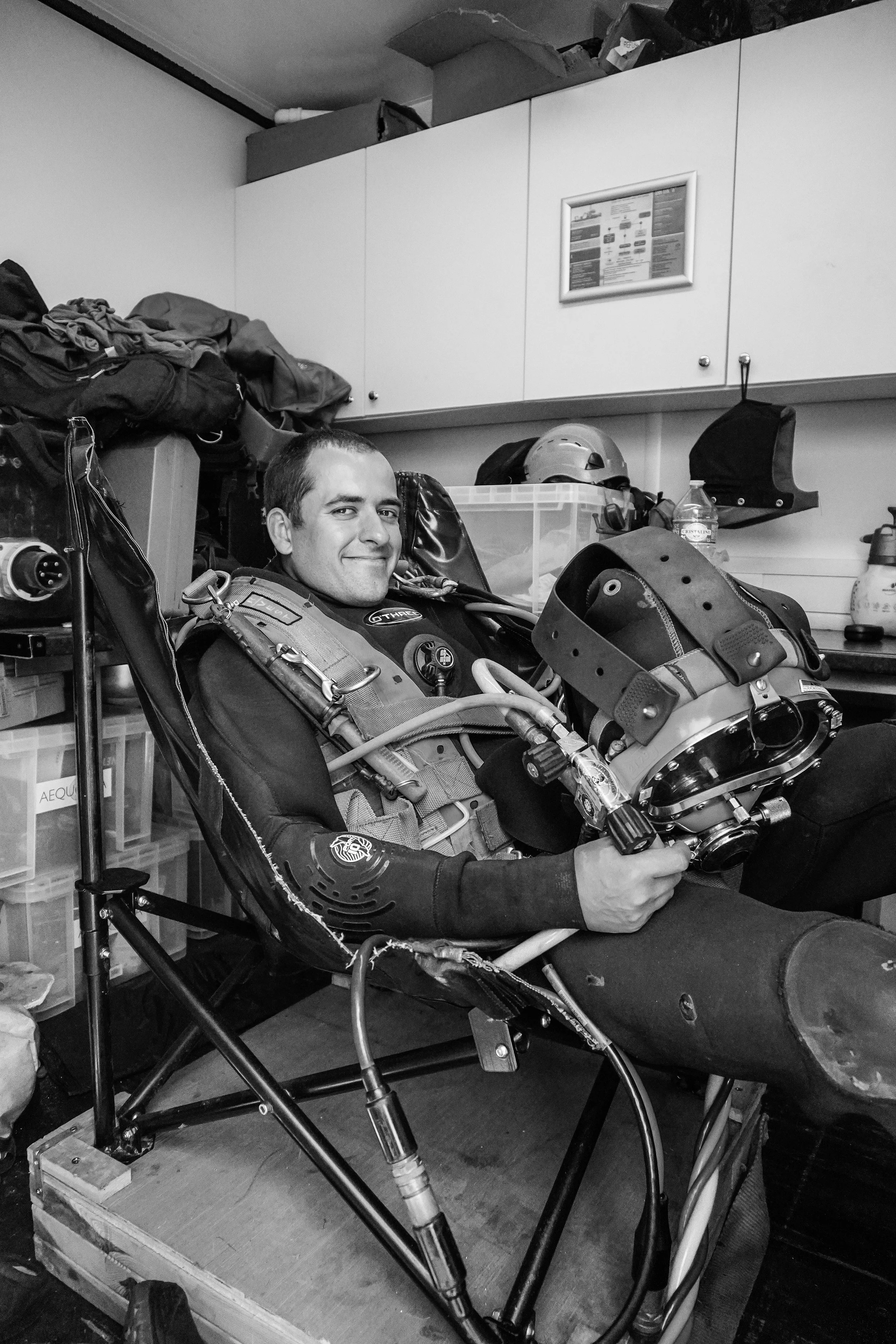 A man sitting in a reclined chair wearing diving equipment, smiling, with various gear and equipment around him in a cluttered room.