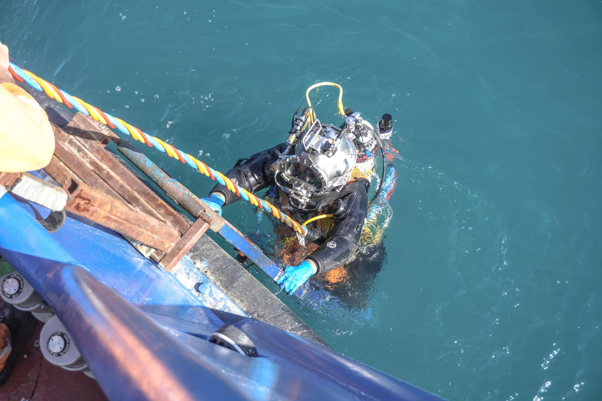 A diver in black drysuit and gloves emerging from water, holding onto a platform with a headset or helmet with attached equipment, with a blue boat and rusted metal structure nearby.