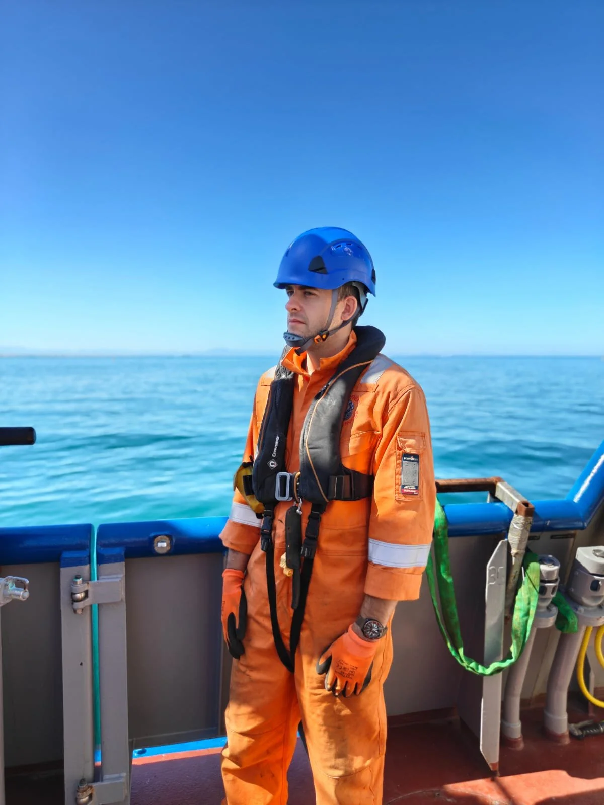 A man dressed in orange safety gear and a blue helmet on a boat, with the ocean and clear sky in the background.