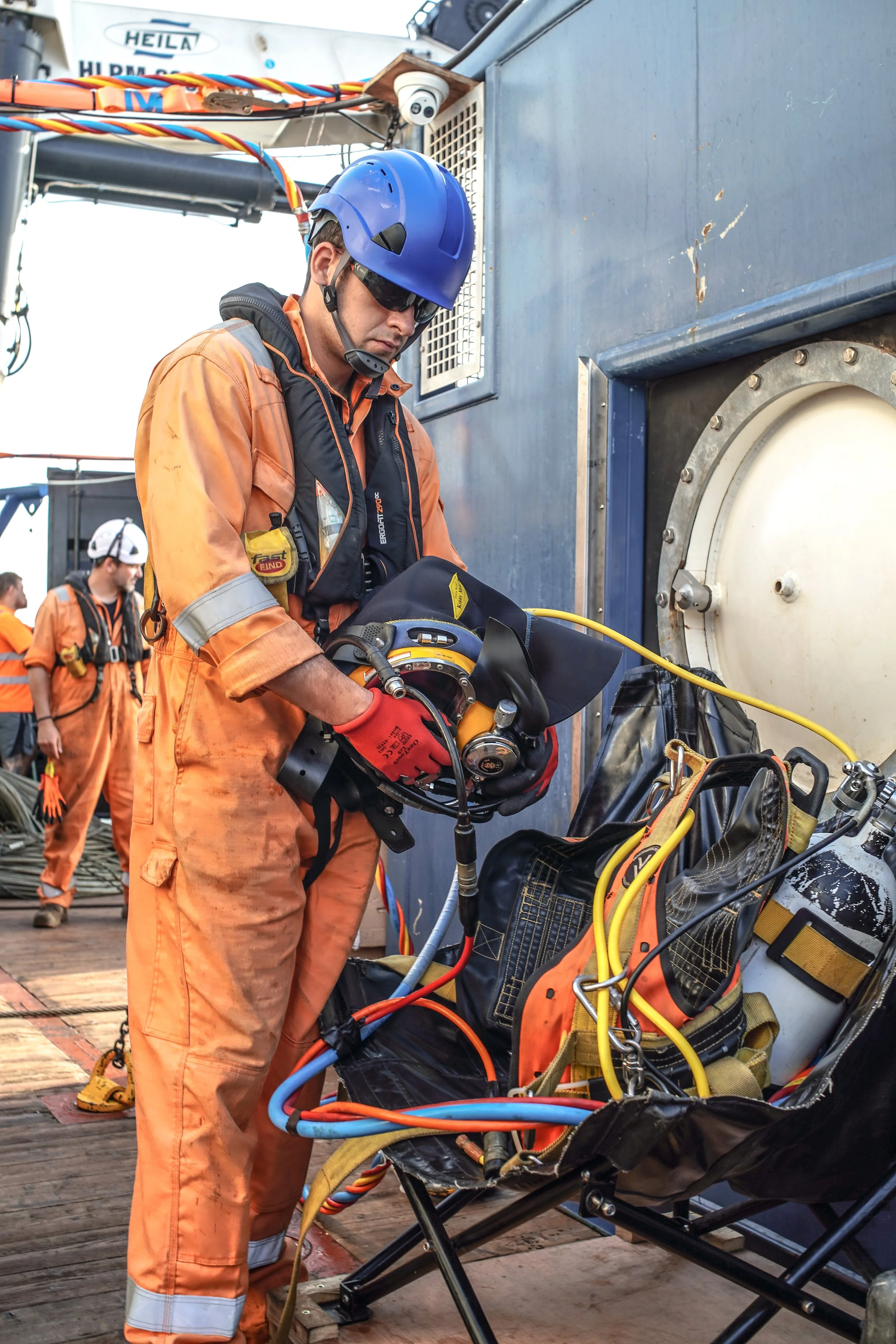 A worker in orange safety gear, a blue helmet, and sunglasses is setting up dive equipment. Two other workers in orange gear and helmets are visible in the background.