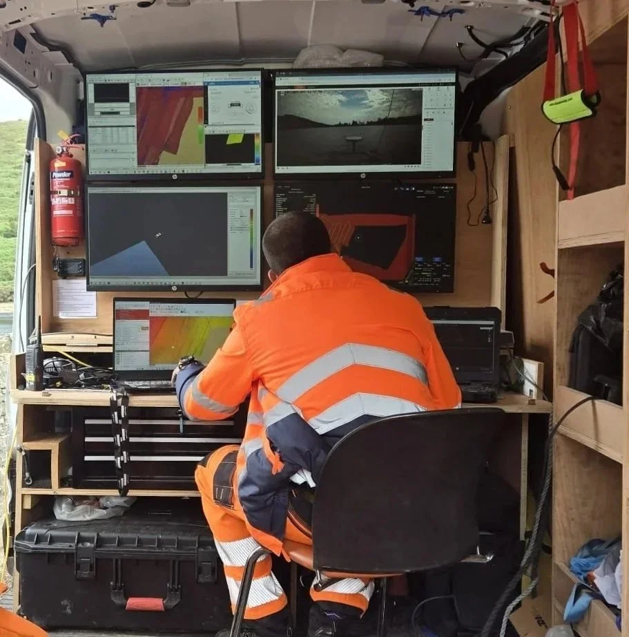 Person in orange safety gear sitting at a desk inside a mobile command or monitoring station with multiple computer screens displaying data and imaging related to surveying.
