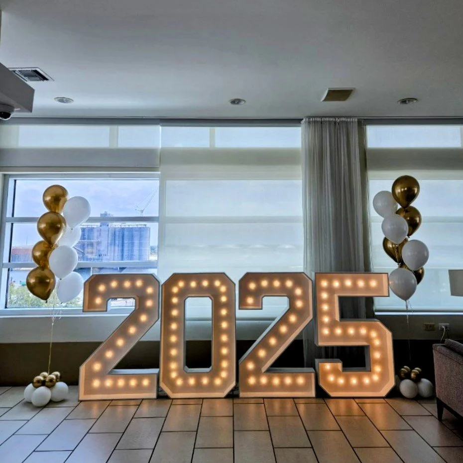 Large illuminated numbers spelling out 2025 with gold and white balloons on either side. Graduation decorations