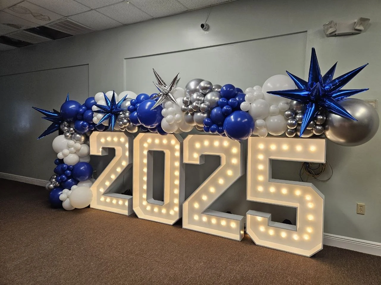 Decorative display for New Year's celebration with illuminated numbers '2025' and blue, white, and silver balloons forming a festive arch.