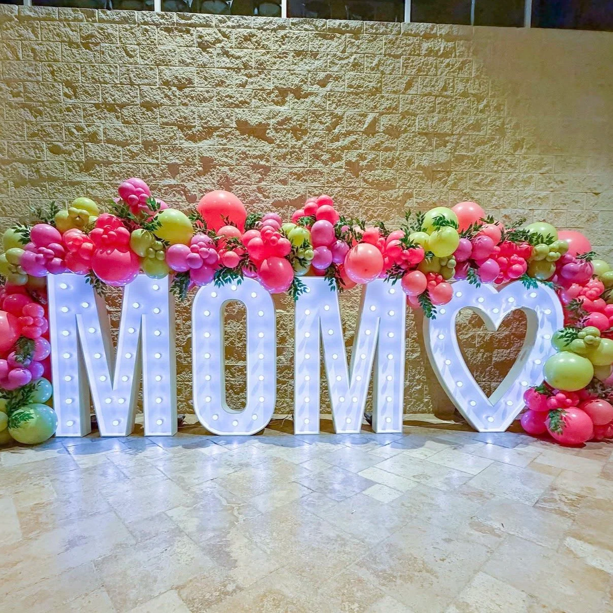 Decorative display spelling 'MOM' with 5 foot large illuminated letters and a large heart marquee surrounded by pink and gold balloons and greenery, set against a textured beige brick wall.