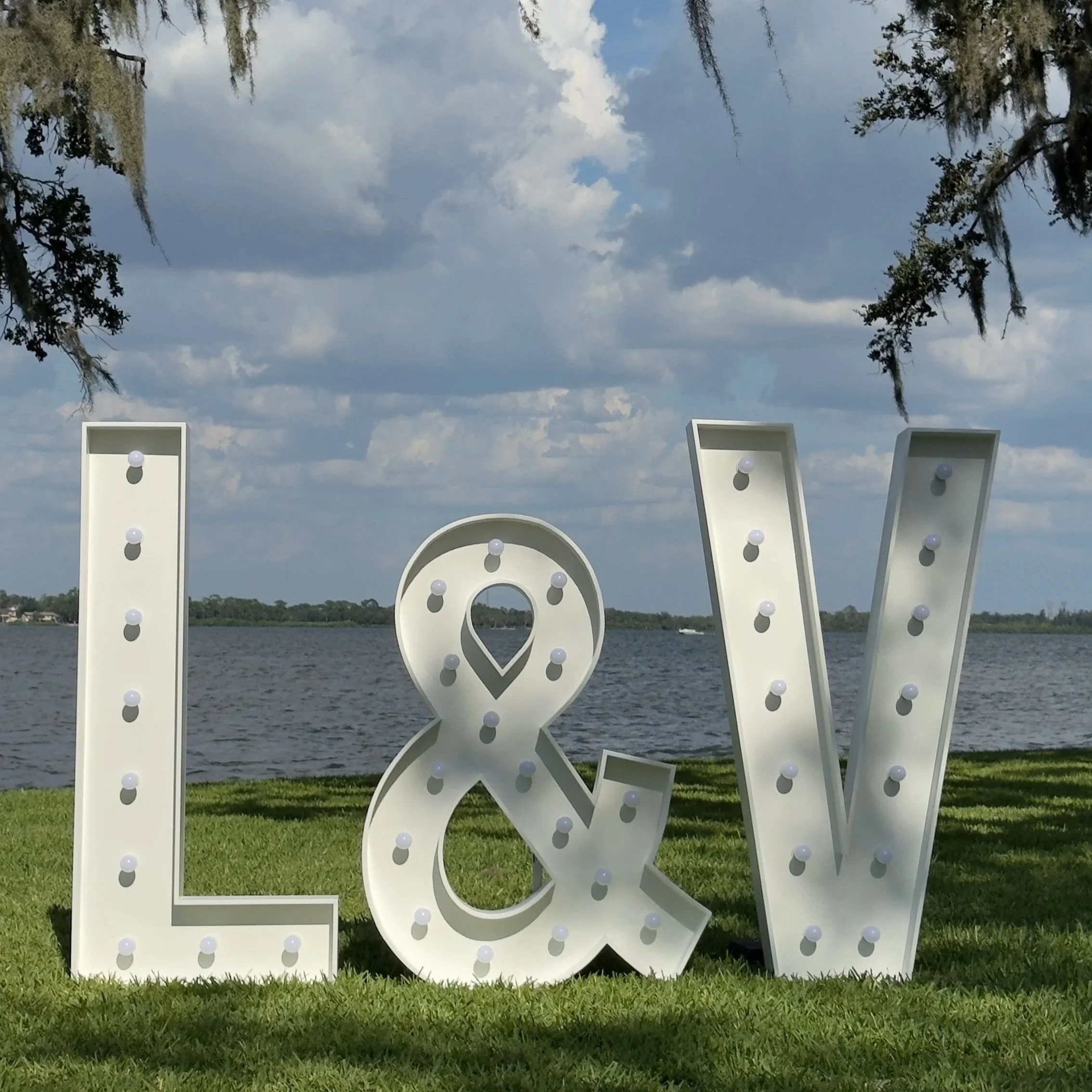 Large five foot white decorative letters L & V with color changing led light bulbs on each letter, positioned outdoors on a grassy area with water and trees in the background and a cloudy sky overhead.