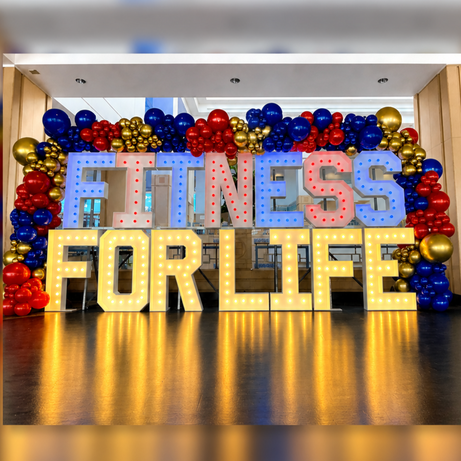 Decorative display with large illuminated letters spelling 'FITNESS FOR LIFE' surrounded by red, blue, and gold balloons.