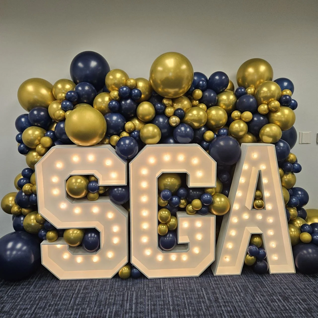Decorative display with large illuminated letters spelling 'SGA' surrounded by a blue and gold balloon garland for a special college school event.