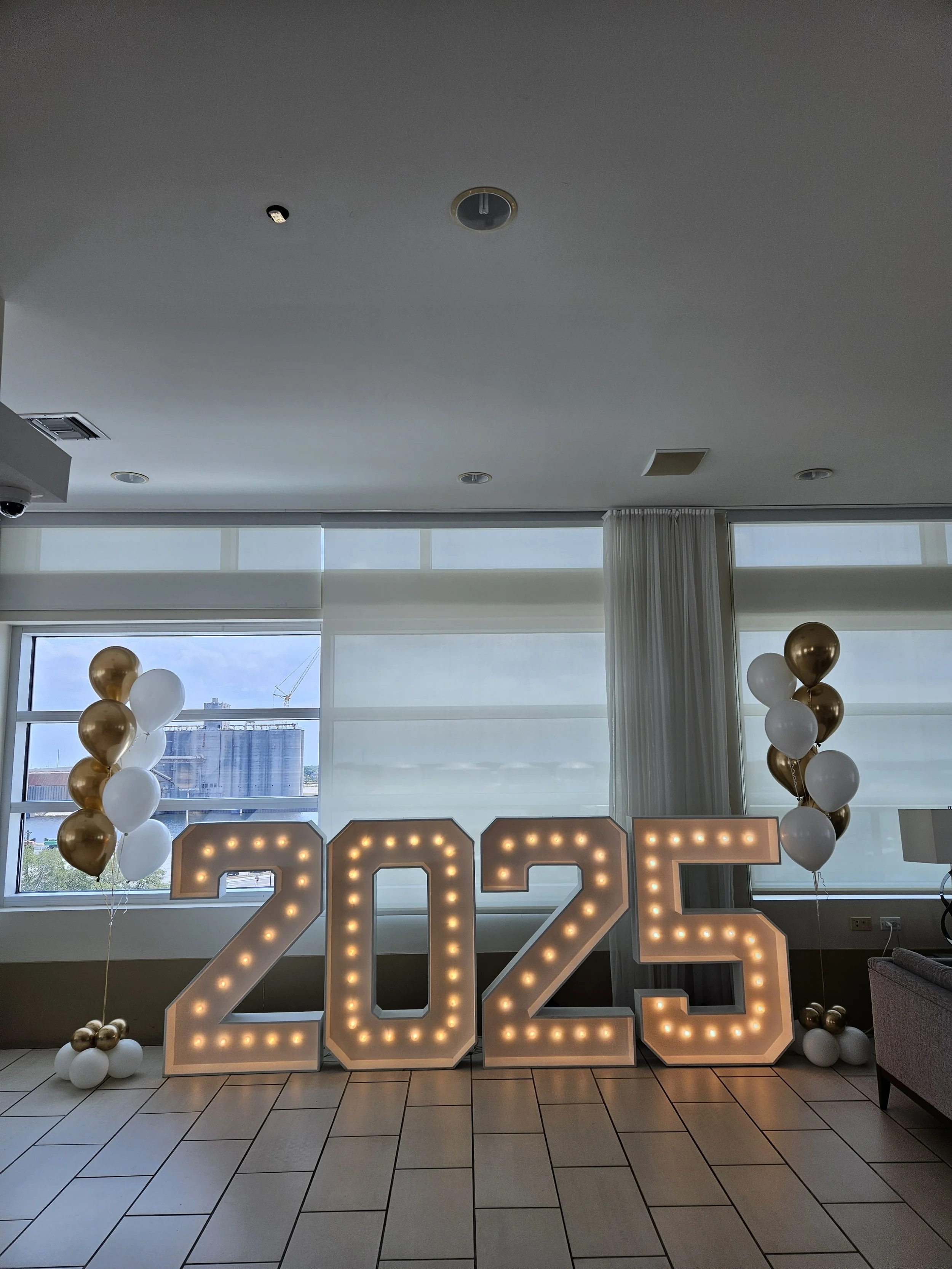 Large illuminated number display showing 2025 with floating balloon bundles on either side. Graduation Decor