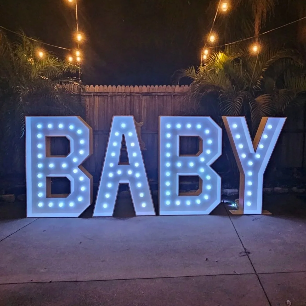 Large illuminated letters spelling 'BABY' with blue lights, set outdoors at night with string lights overhead and palm trees in the background. Great setup for a baby shower.