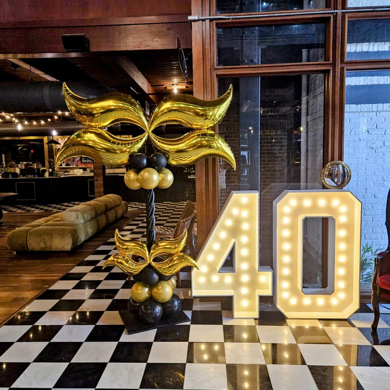 Decorative celebration setup with large illuminated number 40, gold and black balloons, and a black and white checkered floor in a modern indoor venue. Classic 40th birthday event.