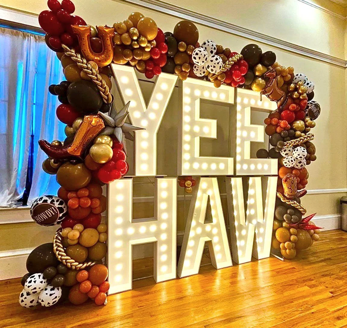 Decorative western display with large illuminated letters spelling "YEE HAW" surrounded by colorful balloons in shades of red, gold, black, and white, with various patterns and metallic finishes, arranged in a festive setting. Prom dance decor