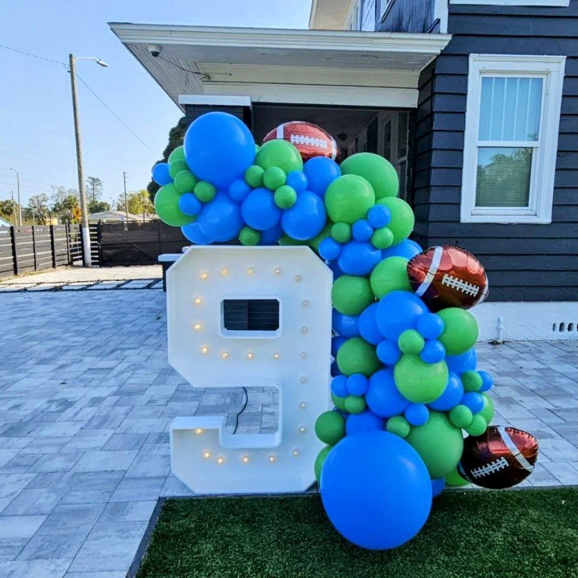 Large white decorative number 9 illuminated with small lights, decorated with green, blue, and a few football-shaped balloons, placed outdoors for a 9th birthday celebration.