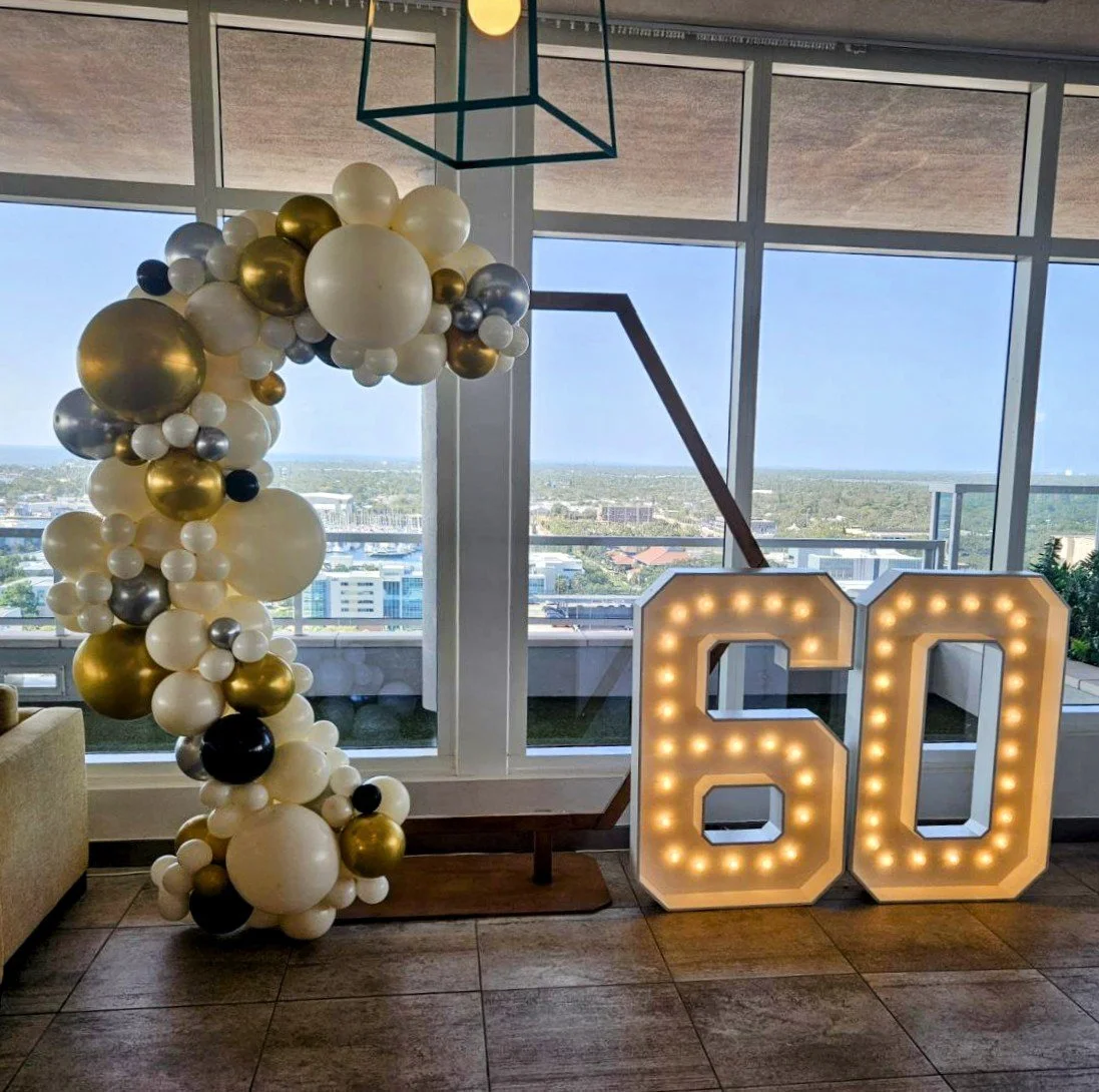 Balloon arch in white, gold, black, and silver colors beside a lighted marquee number 60 sign at a celebration, with large windows and city views in the background. 60th birthday party.