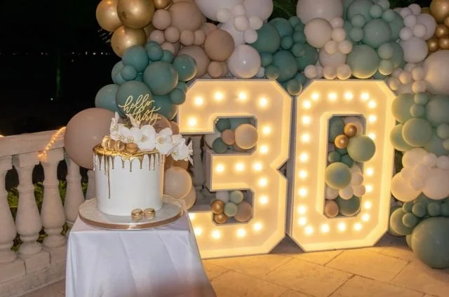 Decorative birthday celebration with a large illuminated '30' and balloons in pastel colors, a white cake with gold accents, and a 'hello my love' cake topper.