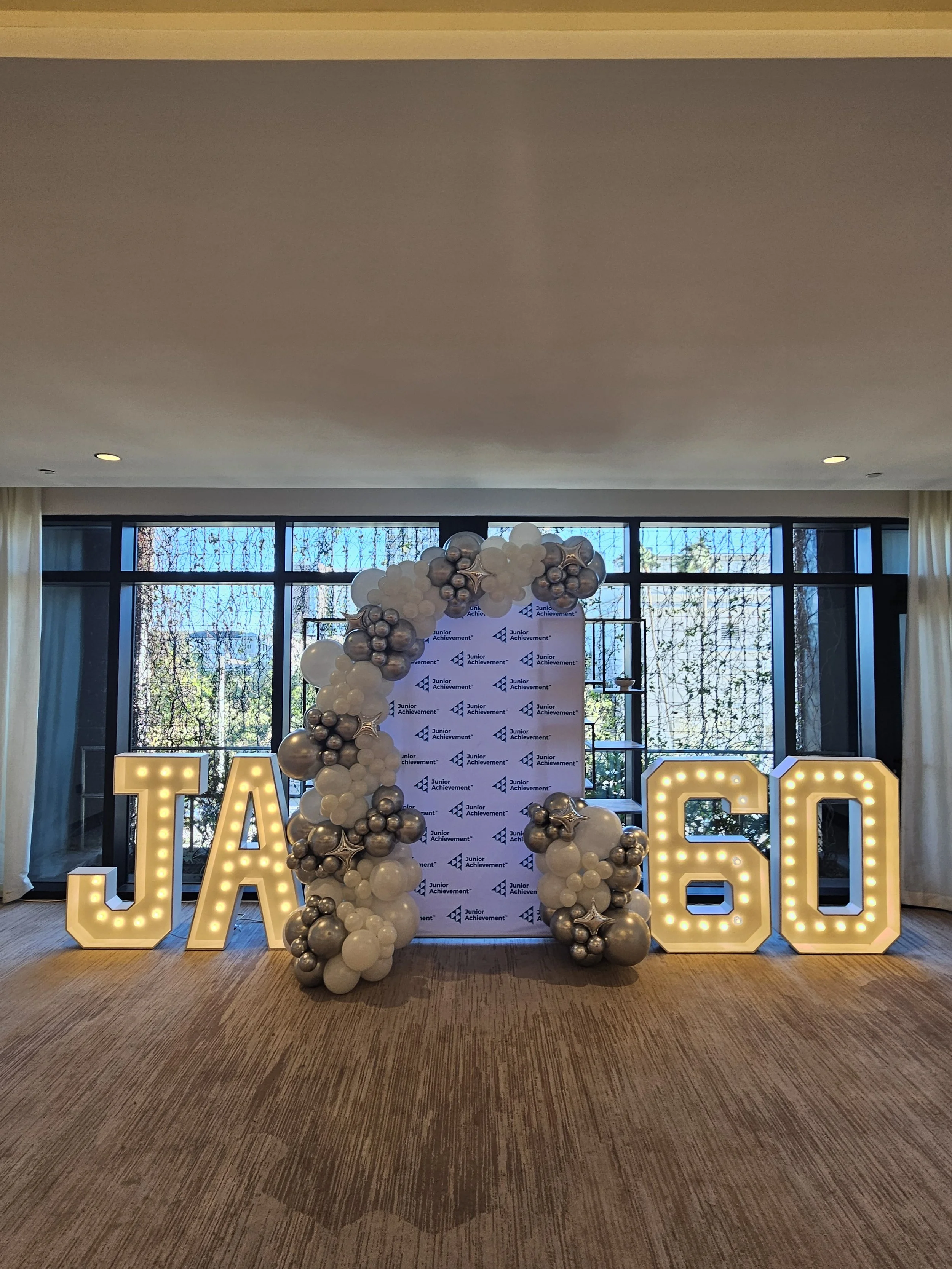 Decorative display with large illuminated letters and numbers, a balloon arrangement with white, silver, and star-shaped balloons, and a backdrop with a repeating logo for Junior Achievement.