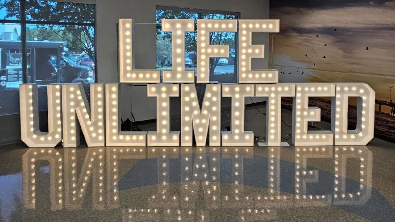 Large illuminated sign reading "LIFE UNLIMITED" with bright bulbs, reflected on a shiny tiled floor. Corporate marquee letter set up
