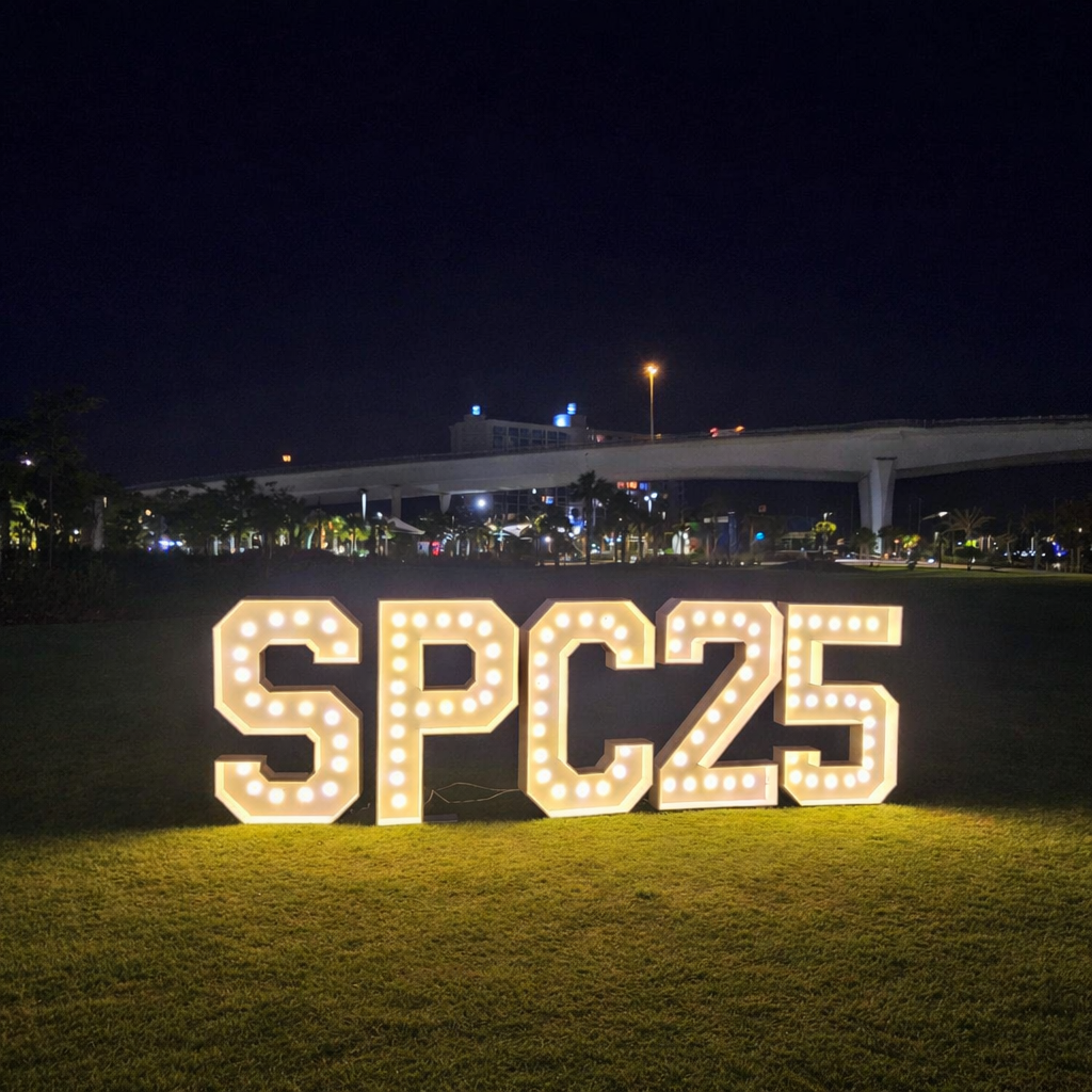 Light-up sign spelling 'SPC25' on a grassy area at night, with a cityscape and elevated highway in the background. St Pete College Graduation decor