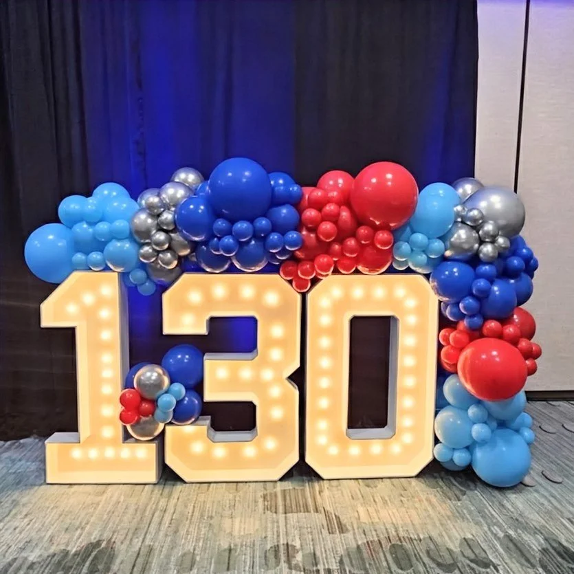 Large illuminated number 130 surrounded by blue, silver, and red balloons. Corporate Anniversary.