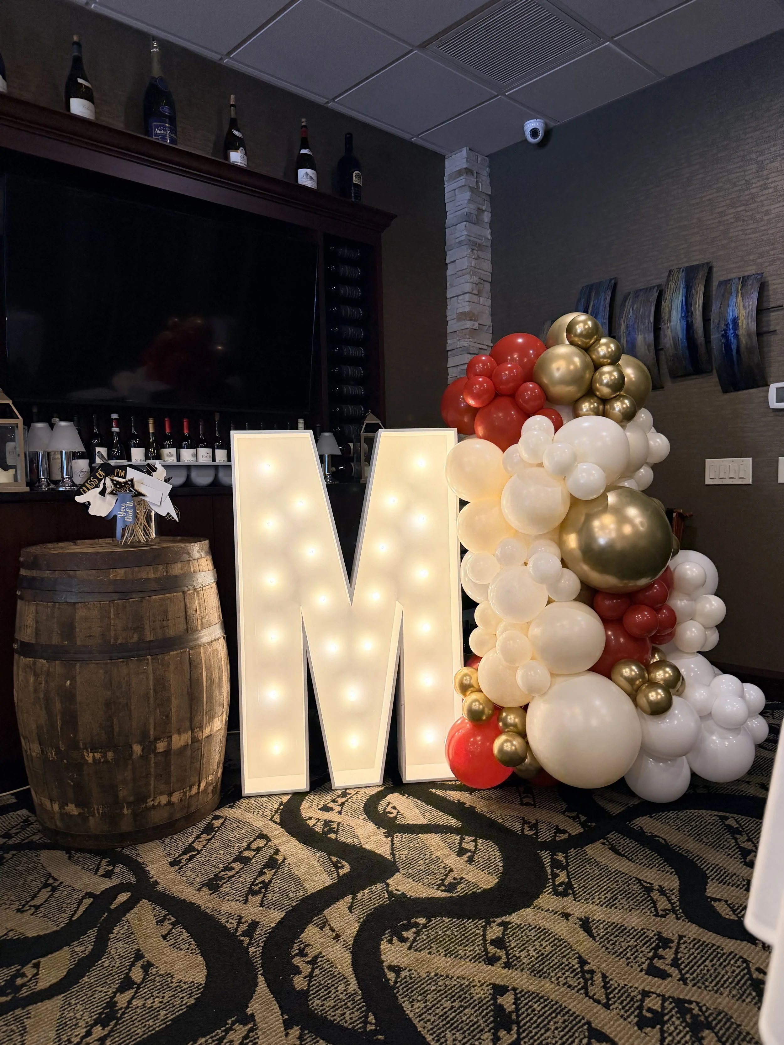A large illuminated letter "M" decoration next to a cluster of red, gold, and white balloons. Graduation marquee and balloon set up