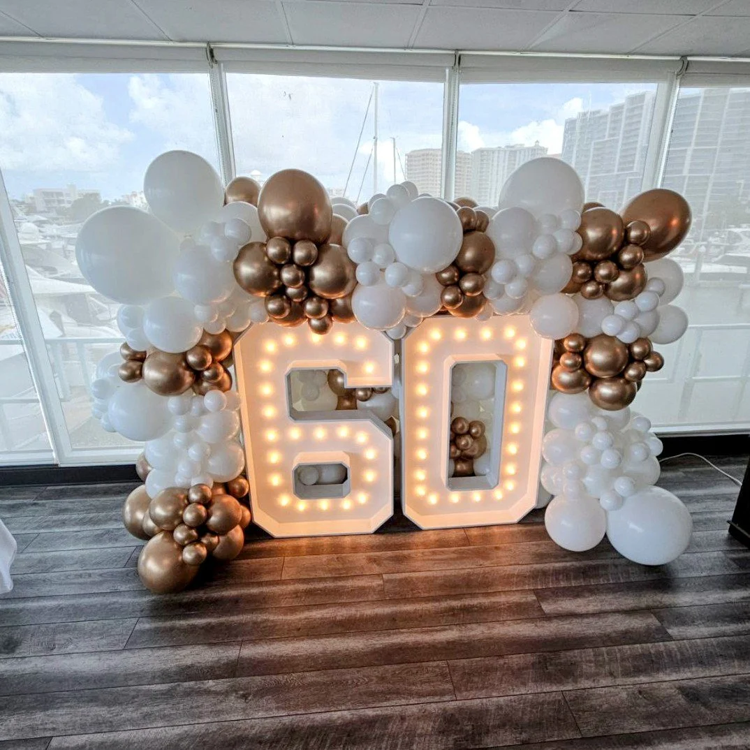 Decorative display with illuminated numbers 60 surrounded by white and metallic bronze balloons at a celebration venue with large windows and city skyline in the background. 60th Birthday balloon and large lit-up letter display