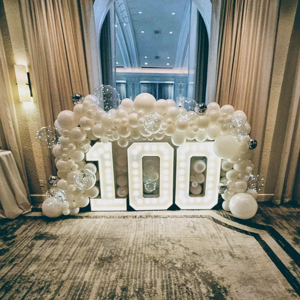 100TH CELEBRATION WITH ALL WHITE AND CLEAR BALLOON GARLAND AROUND MARQUEE NUMBER 100