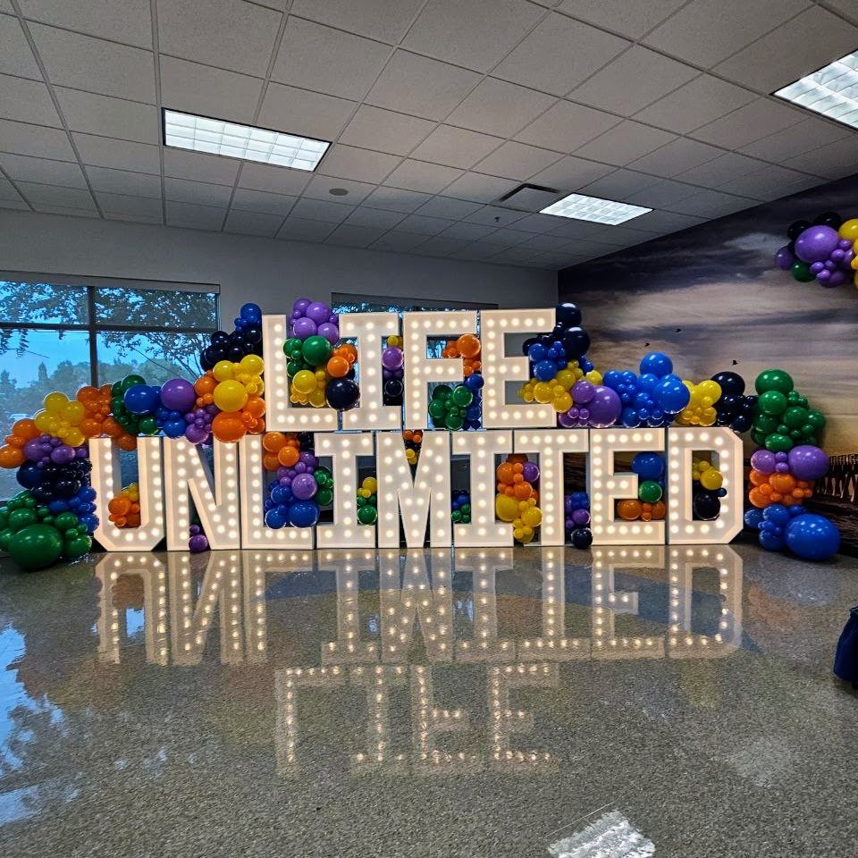 Decorative display with large illuminated letters spelling "LIFE UNLIMITED" surrounded by colorful balloons in purple, yellow, green, black, orange, and blue, placed inside a room with windows and a tiled ceiling.