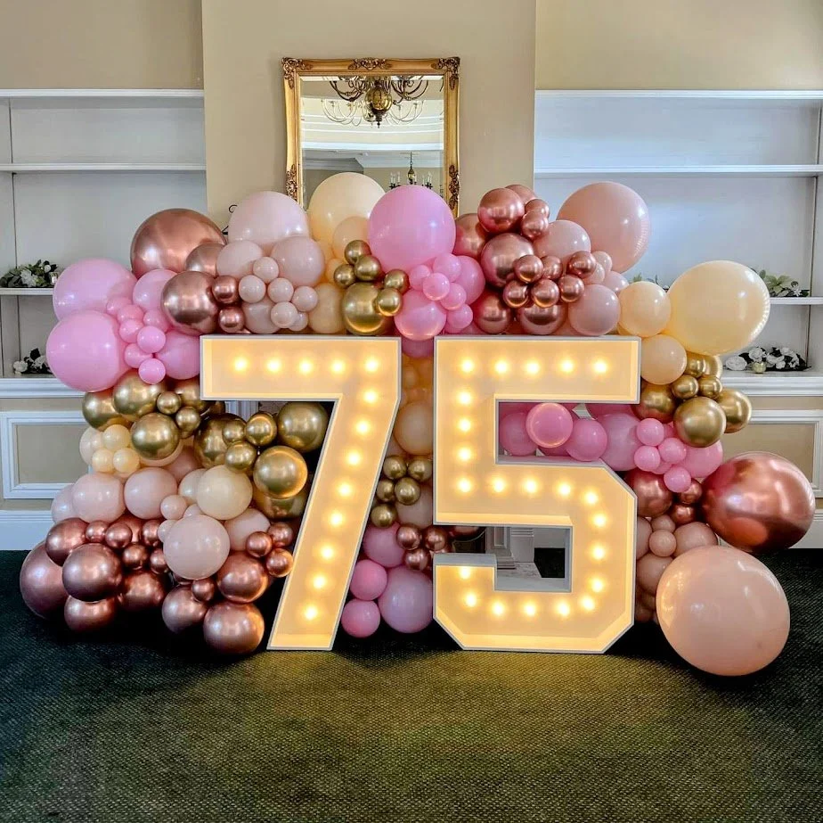 Decorative balloon arrangement with pink, gold, and cream balloons surrounding a large lighted numeral 75. Memorable birthday display