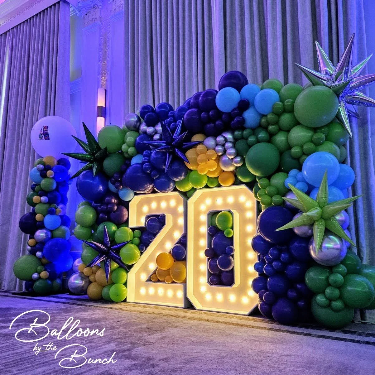 Decorative balloon arrangement with the number 20 in illuminated marquee lights, featuring various green, blue, black, gold, and silver balloons and metallic star-shaped balloons, set against a backdrop of curtains. Bold corporate event set up.