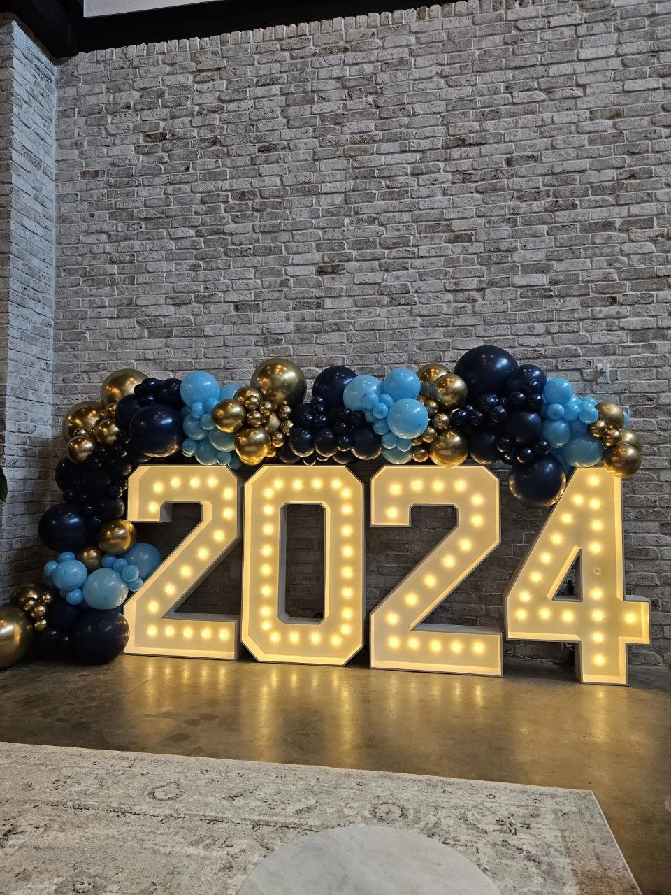 Illuminated number display reading 2024 with black, light blue, and gold balloons above and around it, set against a brick wall. Graduation themed set up