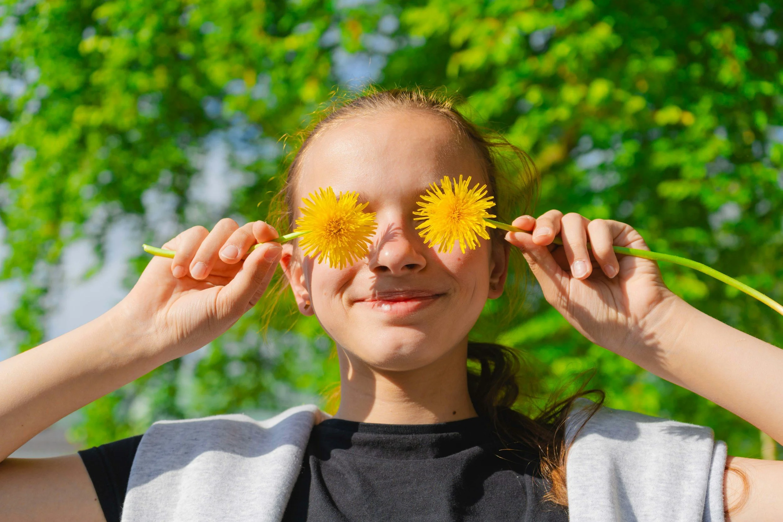 A girl holding yellow dandelions in front of her eyes outdoors with a green leafy background.