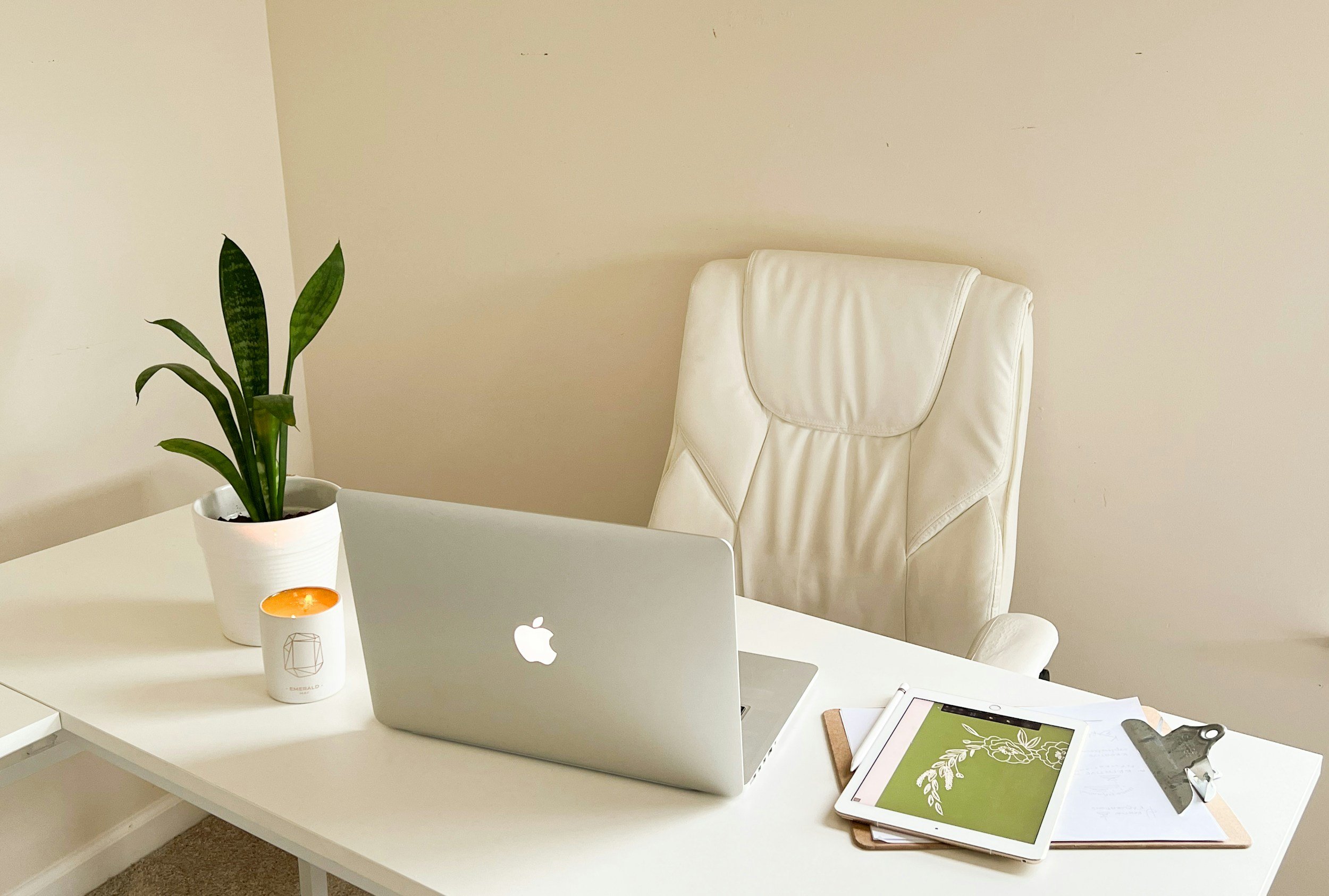 A white desk with a laptop, a white coffee mug, a tablet, and a pad of paper, with a white office chair, a plant, and a candle, in a minimalist workspace.
