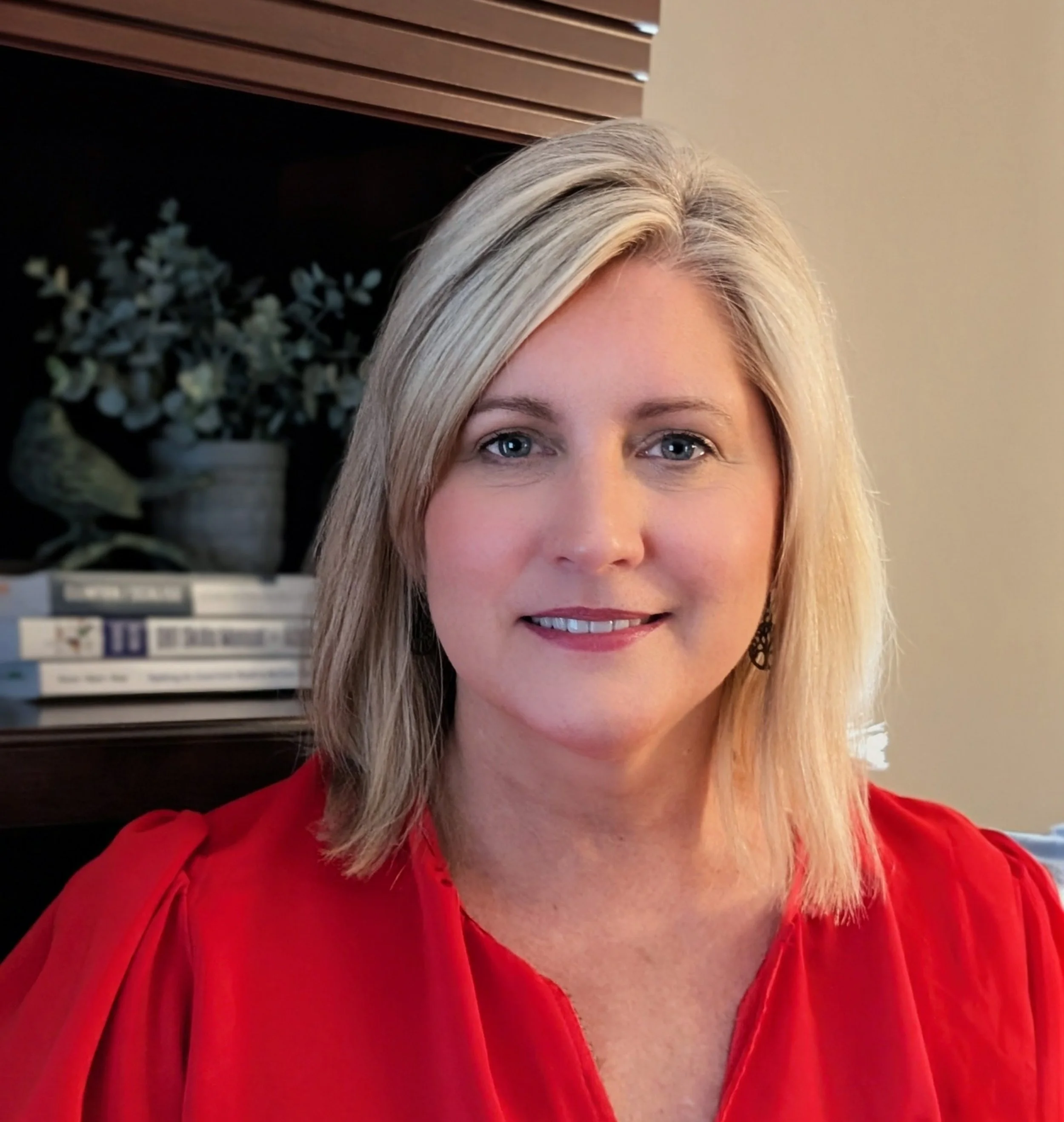 Close-up of a woman with blonde hair wearing a red blouse, showing her face and part of her outfit, with background elements including a potted plant and window blinds.