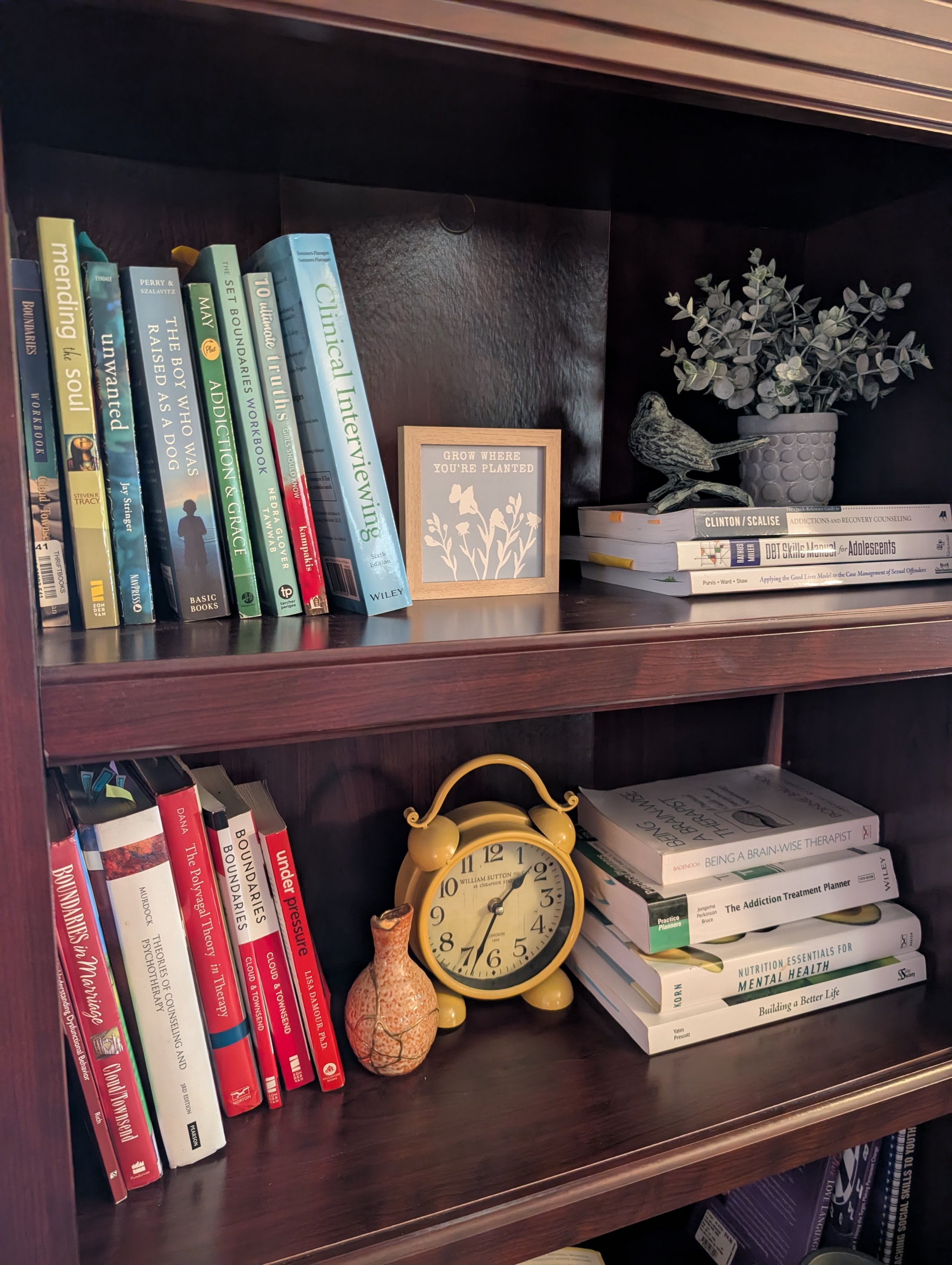 Bookshelf with various books, a yellow clock, a decorative bird, a framed quote, a ceramic vase with eucalyptus, and other books and objects.