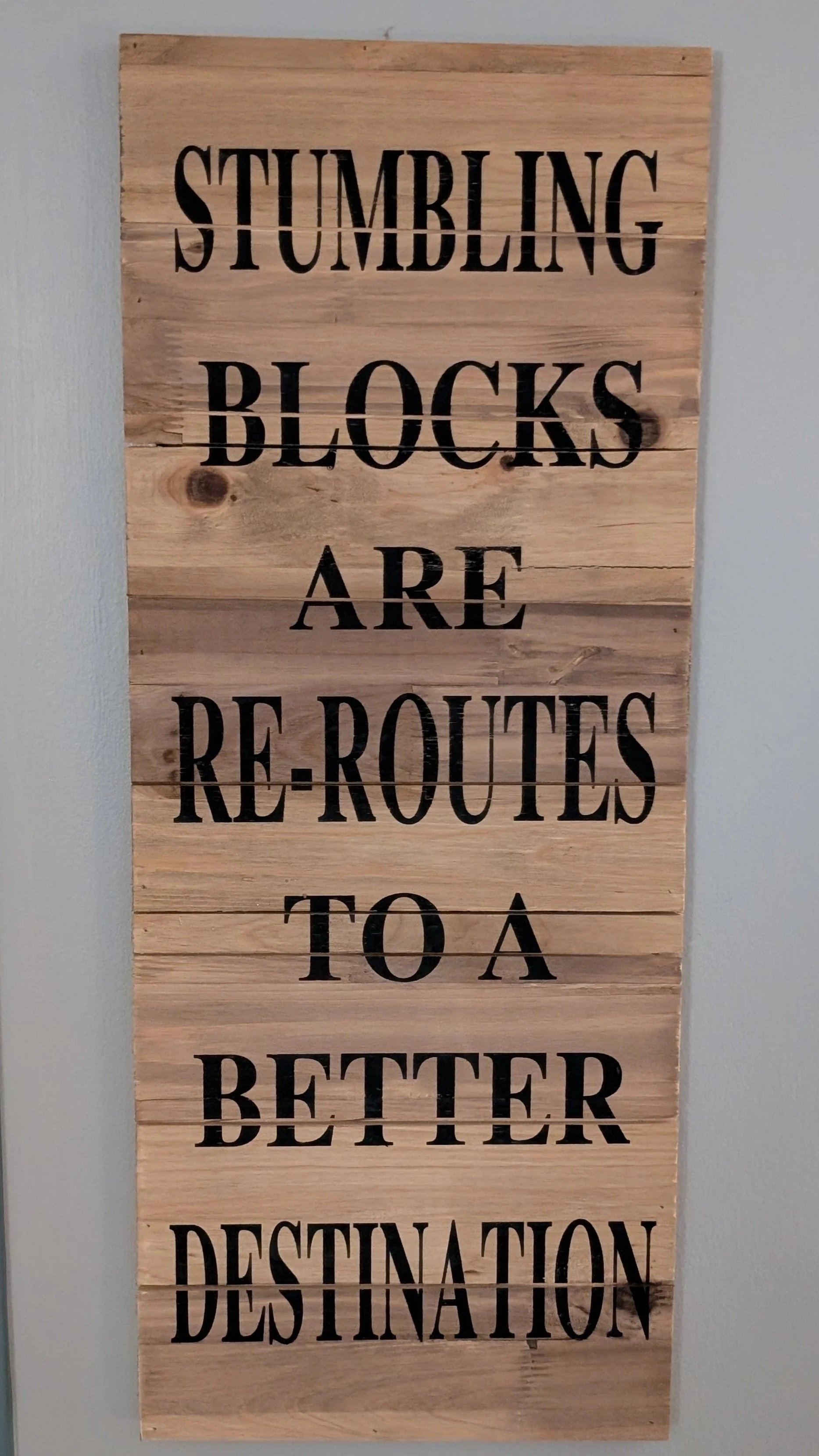 Wooden sign with black stenciled text that reads, "Stumbling Blocks Are Re-routes to a Better Destination."