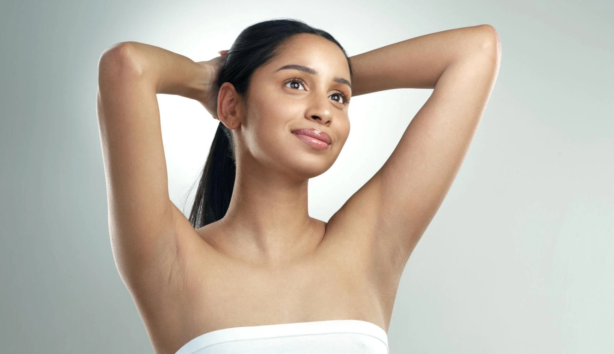 A woman with long dark hair, smiling and with her arms raised behind her head. She has a smooth, clear complexion and is wearing a white strapless top.