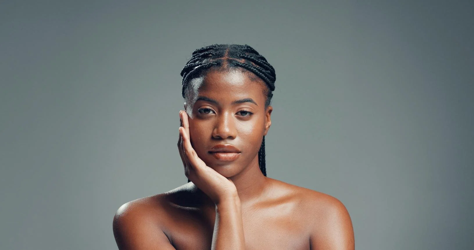Portrait of a young woman with braided hair touching her face against a neutral background.