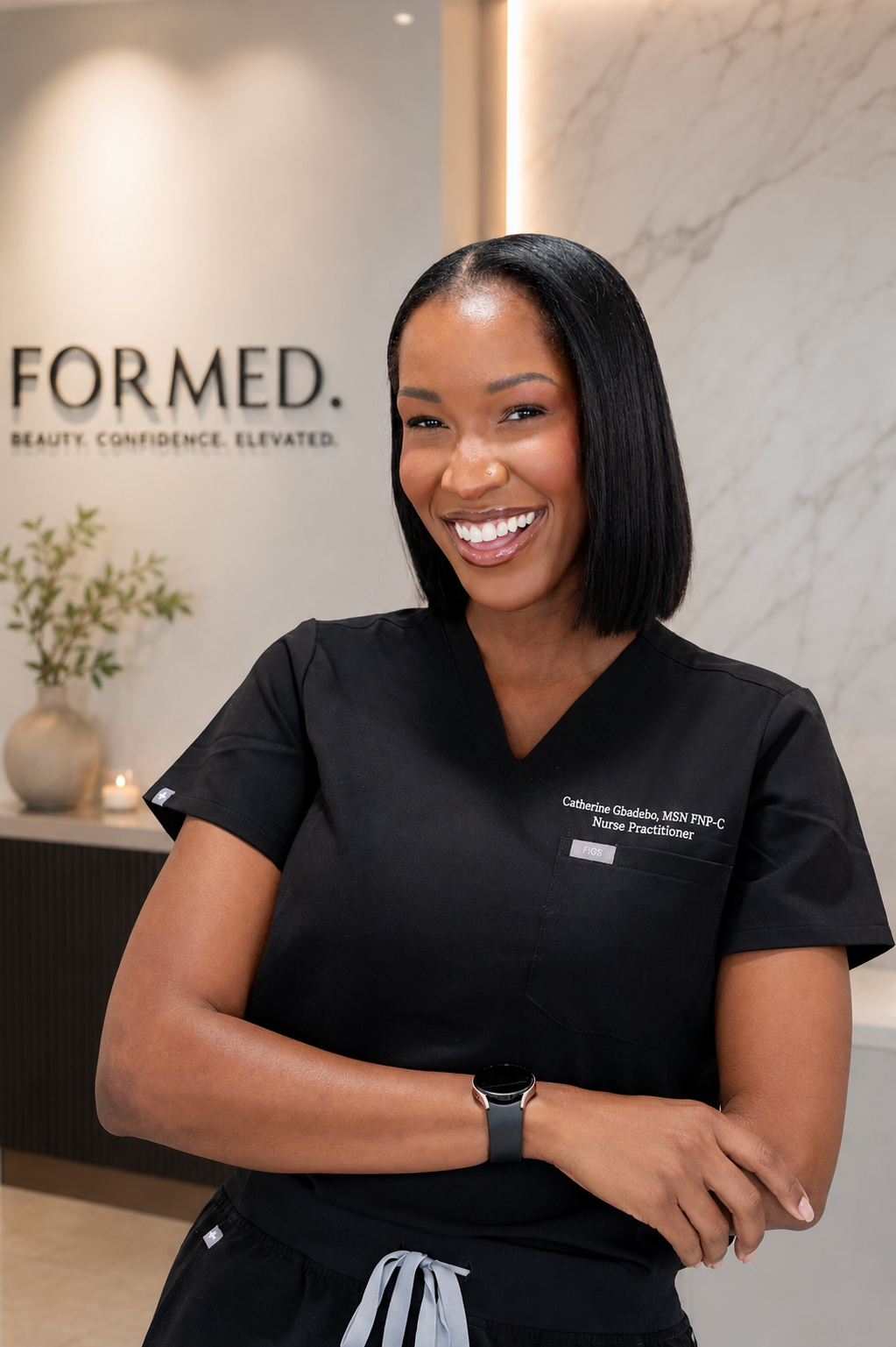 A smiling African American woman wearing black medical scrubs with her name and title, standing in a modern, well-lit reception area with a marble wall and a potted plant in the background.