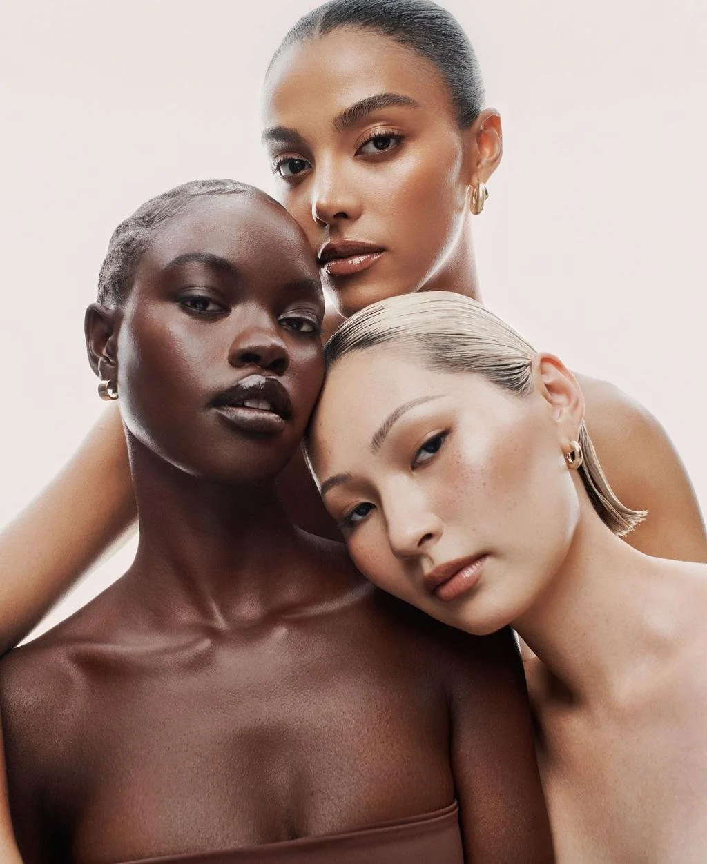 Three diverse women with their heads close together, posing for a portrait against a light background.