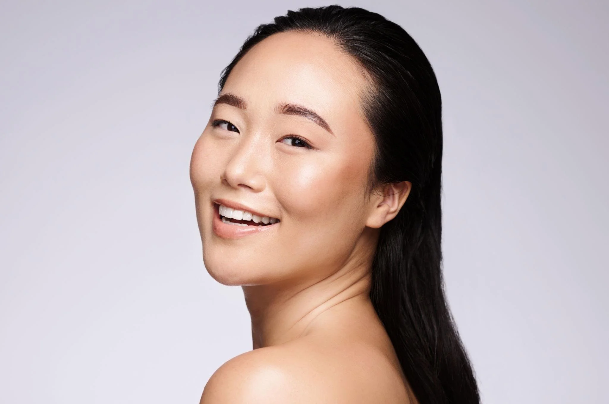 Close-up of a smiling woman with dark hair and natural makeup looking slightly over her shoulder on a plain light background.