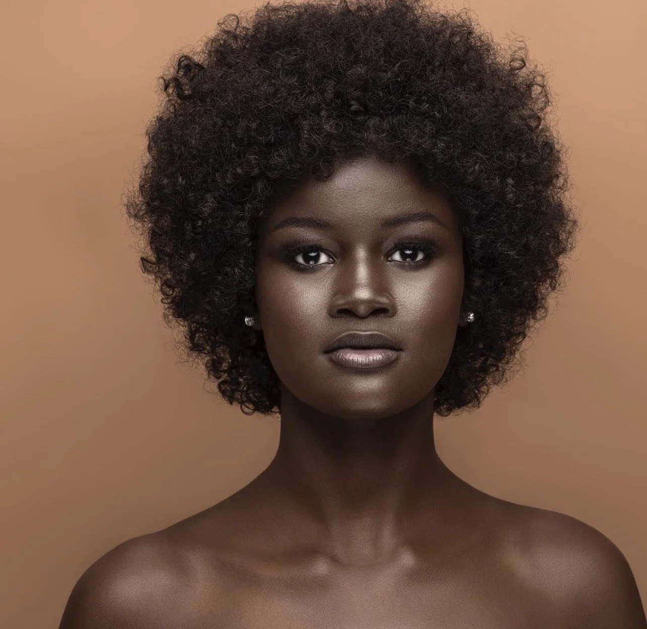 Portrait of a woman with dark skin, short curly black hair, and neutral expression, wearing small earrings, against a beige background.