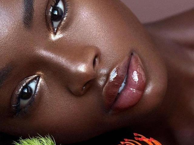 Close-up of a woman's face with glossy lips and shiny skin, lying on a surface with a hint of a colorful object in the corner.