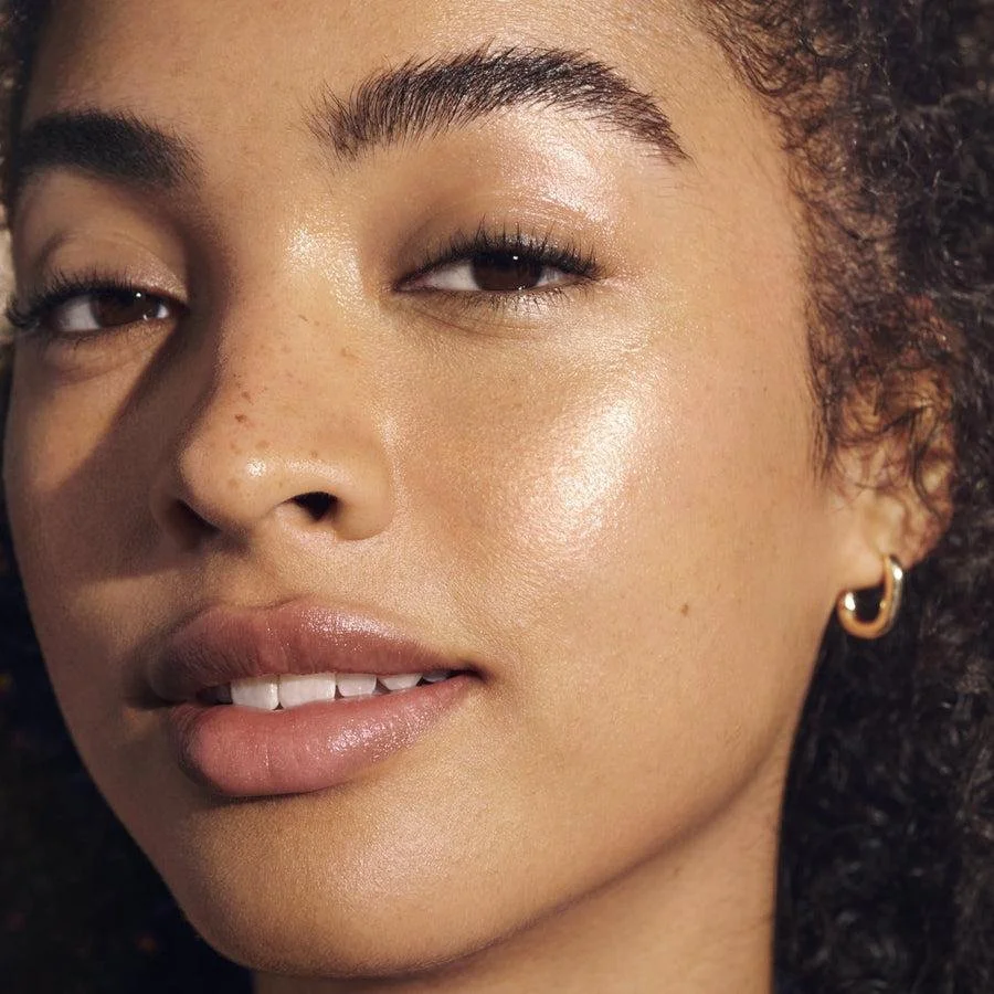 Close-up of a woman with glowing skin, natural makeup, curly hair, and a small hoop earring.